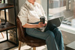 © satura_ - Woman holding coffee cup while working on laptop in sunny cafe. Comfort, routine, and balance between work and relaxation in lifestyle.