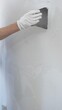 © volha_r - Construction worker wearing gloves and blue overalls is using a putty knife to apply plaster on a white wall, performing home renovation and improvement, close-up of hand