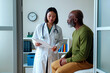© pressmaster - Asian young adult female doctor discussing medical results with Black middle aged man sitting on examination table in modern clinic office, both focused on clipboard information