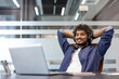© Tetiana - Smiling young Indian man wearing headphones sits at a desk in the office, hands behind his head, and looks contentedly at the laptop screen