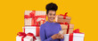 © Prostock-studio - A happy woman with curly hair smiles as she stands near a pile of beautifully wrapped gift boxes. She is holding a smartphone and seems excited. The bright yellow background adds warmth to the scene.