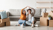 © Prostock-studio - Two joyful millennial Asian individuals sit on the floor among carton boxes in their new home, making a house roof gesture. It is a special moment on their moving day filled with excitement.