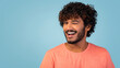 © Prostock-studio - A cheerful young eastern man with curly hair and a beard poses in a stylish orange shirt. He winks and smiles at the camera in front of a blue studio background, capturing a fun moment.