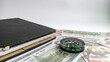 © Gabie - The compass is placed on top of a spread of hundred dollar bills and the left side of a black notebook, symbolizing time, wealth, and guidance in financial decisions and investment strategies.