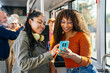 © Koldo_Studio - Two smiling multi ethnic women friends are sharing a smartphone while traveling on a city public bus, representing urban commute and connection