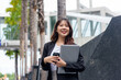 © sorapop - Young Asian business woman, busy professional, wears suit, stands on street, carrying laptop, coffee for work, exudes confidence, smiles brightly in modern urban environment.