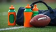 © Vadym - American football rests on bright green grass field near white sideline. Three vibrant orange, green water bottles stand close. Dark sport bag also nearby. Items show summer sport training,