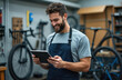 © Vadym - Young handsome bearded man mechanic smiles, looking at digital tablet in bicycle repair shop. Wears protective apron. Professional service person checks orders, inventory, maintenance schedules using