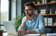 © Vadym - Young man works on laptop in modern office space. Wears glasses, blue shirt, looks focused at screen. Guy thinks about new business project, strategy, future plan. Busy male worker sitting at desk,