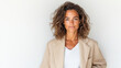 © siraphol - Woman with curly hair wearing beige blazer and white top stands confidently against plain background. Her expression is calm and poised