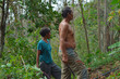 © agratitudesign - A shirtless father and his wife in a blue shirt stand side by side observing the contrast between cleared and overgrown areas of their tropical hillside farmland.
