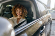 © SHOTPRIME STUDIO - Smiling woman driving a car with curly hair wearing a white blouse, enjoying a sunny day in an urban environment with modern buildings in the background.