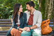 © Srdjan - Young couple cuddling on a park bench, smiling and leaning in for a kiss, sharing an affectionate embrace in sunny green nature casual outfits, warm connection, romantic date outdoors