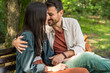 © Srdjan - Young couple cuddling on a park bench, smiling and leaning in for a kiss, sharing an affectionate embrace in sunny green nature casual outfits, warm connection, romantic date outdoors