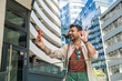 © Srdjan - Young traveler laughing while taking a selfie in a modern business district cheerful millennial man with backpack capturing urban architecture and city vibes on smartphone during a sunny day