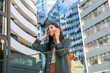 © Srdjan - Smiling young woman on phone call in modern business district cheerful traveler with backpack, chatting on smartphone amid sleek urban architecture and reflective office towers on a sunny day