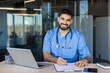 © Liubomir - Young male doctor or medical student in blue scrubs and stethoscope smiling at the camera while writing notes on paper, sitting at a desk with a laptop in a modern office