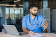 © Liubomir - Male healthcare professional in blue scrubs and stethoscope reviewing patient information on a digital tablet while simultaneously writing notes on a clipboard, at an office desk with a laptop