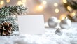 © Igor_Tichonow - White blank holiday card standing on snow with pine cone and silver ornaments illuminated by warm festive lights creating cozy christmas mood