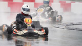 Kids' karting. Two kids' racing cars turn on a wet go-kart track. The karts' wheels spray water, and the cars are reflected in the puddles.