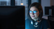© Atmospheres - Focused female programmer wearing glasses works on a computer late at night in a dark office, smiling with satisfaction.