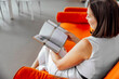 © Айман Дайрабаева - young Hispanic woman with short brown hair sits on an orange couch in a library, reading a book. Bookshelves filled with books are visible in the background.