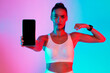 © Prostock-studio - A fit woman stands in a colorful space with bright blue and pink lights. She holds a smartphone towards the camera and flexes her arm to display her strength.