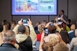 © Anton Gvozdikov - Crowd watches speaker on stage at conference while attendees record slides with smartphones and cameras creating dynamic social video moment