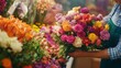© Artsaba Family - A colorful array of fresh flowers including carnations, dahlias, and roses in various shades, held by a person wearing an apron at a bustling flower market