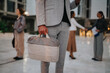 © qunica.com - A business professional in a grey check suit carries a beige bag in a contemporary city plaza, with colleagues meeting and greeting in the background.