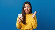 © Atmospheres - Surprised woman with a smartphone celebrates good news. Excited Asian girl reacts to an amazing online offer or win against a blue background.