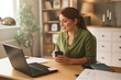 © Stockphotodirectors - A woman sits at a wooden desk in a warm home office, engaging in a video chat with a laptop. She holds a cup of coffee and appears cheerful, surrounded by greenery and soft lighting.