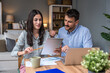 © Srdjan - Couple reviewing household budget at home woman with coffee and tablet, man focused on laptop; paperwork on table in cozy living room, finances, bills, savings, remote work.