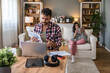 © Srdjan - Man reviews bills at a laptop while his partner relaxes with a tablet on the sofa in a cozy living room. Papers, coffee and tech on the table. Concept: home budgeting, monthly expenses