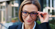 © serg - Close up portrait smiling face business woman wearing stylish glasses looking at camera posing outdoors at modern office building on background. Female manager employer entrepreneur. Business concept.
