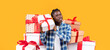 © Prostock-studio - A young man stands holding a gift box, smiling as he poses in front of a tall stack of colorful presents. The cheerful scene has a bright yellow background highlighting the festive atmosphere.