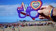 © Canary4stock - Person wearing rainbow wristband holds novelty pride glasses with heart logo above crowded sunny Maspalomas beach as diverse crowd enjoys summer LGBTQ+ celebration and festival