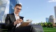 © photostockatinat - An ambitious person in a suit is deeply engrossed in his phone, holding a book, set against a backdrop of the modern buildings