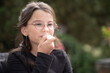 © PeterPike - Dark-haired girl in glasses and long sleeve sitting outdoors, applying lipstick on a blurred natural background, soft daylight, copy space. Concept of growing up, curiosity, and self-expression.