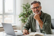 © ArtMajestic - Caucasian man with grey hair and beard wearing glasses and green shirt smiling and looking at camera at desk with laptop and notebook. Remote work and successful business lifestyle, with copy space