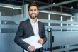 © Liubomir - Confident businessman smiling at camera, standing in a contemporary office environment, holding a laptop, representing professionalism, success, and corporate leadership