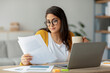 © Prostock-studio - Concentrated arab businesswoman sits at her home office desk, reading papers while holding a cup of hot beverage. She is engaged in her work in a cozy living room setting.