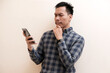 © Nurma - Southeast Asian man wearing flannel shirt showing thinking gesture with his eyes looking smartphone on hand, on white isolated background.