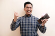 © Nurma - Southeast Asian man smiling wearing flannel shirt holding leather wallet with hand give thumbs up, on white isolated background.