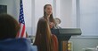 © Framestock - American Teenage Girl Delivers Expressive Speech in Front of Classmates, Gesturing Confidently With US Flag. Concept Civic Education, Confident Communication, Active Student Participation. Dolly Shot.