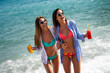© NDABCREATIVITY - Two happy women toasting colorful beach drinks under the sun, enjoying tropical vibes and friendship