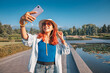 © EdNurg - Young woman taking selfie with smartphone in a park with pond and mountains in background during summer sunny day