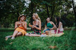 © qunica.com - A group of four women share a relaxed picnic on a grassy park, chatting and laughing while enjoying pastries, a wicker basket, and the warm outdoor ambiance.