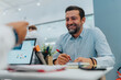 © qunica.com - Smiling bearded man works at a bright, modern office desk, writing with a red marker beside a laptop and charts. The scene conveys teamwork, planning, and a productive multicultural business setting.