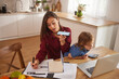 © Stockphotodirectors - A woman talks on the phone while taking notes, seated at a kitchen table. A child plays nearby with a calculator, creating a busy family atmosphere.
